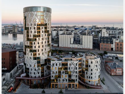 Gesamtansicht des Ensembles in städtischer Situation: weiträumige Außenaufnahme bei Dämmerung zeigt den runden Turm und die niedrigeren, organisch gegliederten Baukörper davor, Hafenanlagen am Horizont; Straße und Nebengebäude im Vordergrund.