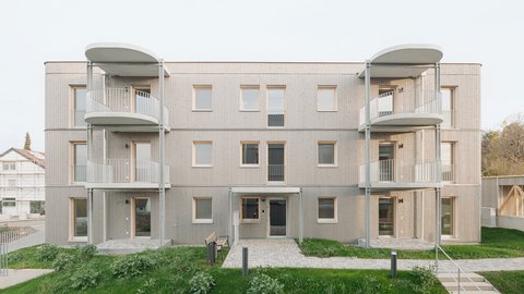 Dreigeschossiges Wohnhaus mit heller Holzfassade und zwei übereinanderliegenden Balkontürmen, davor Grünfläche und gepflasterter Weg.