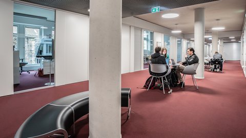 Offener Lernbereich mit mehreren Personen an Tischen und Stühlen auf rotem Teppichboden in modernem Gebäude mit weißen Säulen