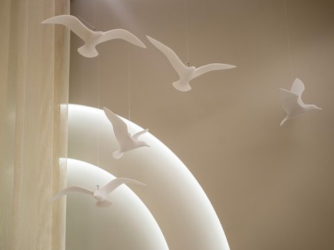 "Detailaufnahme von hängenden weißen Vogelfiguren gegen einen hellen beige-braunen Hintergrund. Fünf stilisierte Vogelskulpturen in verschiedenen Flugposen, von Metallstangen aufgehängt. Links teilweise sichtbare beige Wandverkleidung. Die Objekte werfen Schatten und erzeugen einen eleganten, schwerelosen Eindruck."