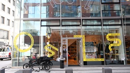 Straßenansicht eines Glasgebäudes mit geöffnetem Eingang und Ausstellung im Erdgeschoss.