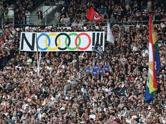 Ein voll besetztes Fußballstadion mit Fans auf den Rängen. In der Mitte wird ein großes Banner mit der Aufschrift „NOOOO!!“ und den olympischen Ringen hochgehalten, daneben sind verschiedene Fahnen zu sehen.