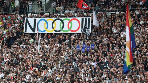 Ein voll besetztes Fußballstadion mit Fans auf den Rängen. In der Mitte wird ein großes Banner mit der Aufschrift „NOOOO!!“ und den olympischen Ringen hochgehalten, daneben sind verschiedene Fahnen zu sehen.