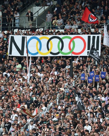 Ein voll besetztes Fußballstadion mit Fans auf den Rängen. In der Mitte wird ein großes Banner mit der Aufschrift „NOOOO!!“ und den olympischen Ringen hochgehalten, daneben sind verschiedene Fahnen zu sehen.