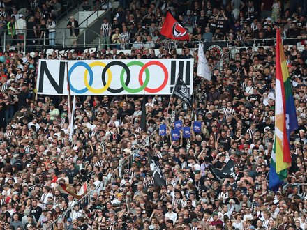 Ein voll besetztes Fußballstadion mit Fans auf den Rängen. In der Mitte wird ein großes Banner mit der Aufschrift „NOOOO!!“ und den olympischen Ringen hochgehalten, daneben sind verschiedene Fahnen zu sehen.