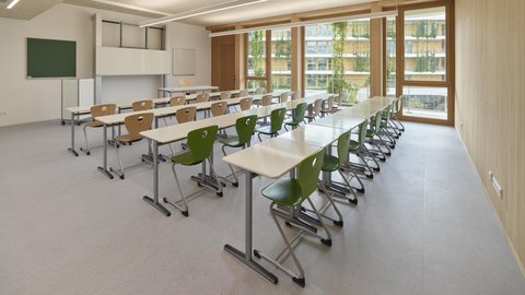 Leerer Klassenraum mit Reihen von Tischen und grünen sowie braunen Stühlen vor großen Fenstern und einer Tafel an der Wand