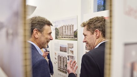 Seitliche Perspektive durch einen schmalen Rahmen. Im Hintergrund Tafeln mit Fotos eines mehrgeschossigen Wohnbaus und erklärenden Texten. Lockere Gesprächssituation in einer Ausstellungsumgebung.