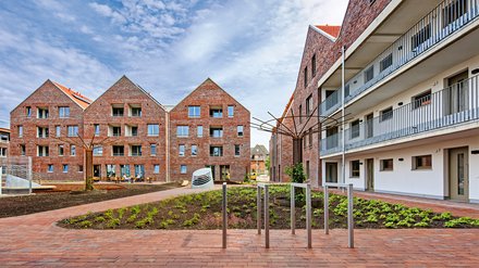 Innenhof mit Wohngebäuden: Blick auf bepflanzte Beete und gepflasterten Platz, links und im Hintergrund rote Backsteinhäuser mit Satteldächern, rechts modernes Wohngebäude mit Balkonen und Außengängen, in der Mitte eine Metallskulptur.