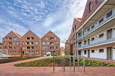 Innenhof mit Wohngebäuden: Blick auf bepflanzte Beete und gepflasterten Platz, links und im Hintergrund rote Backsteinhäuser mit Satteldächern, rechts modernes Wohngebäude mit Balkonen und Außengängen, in der Mitte eine Metallskulptur.