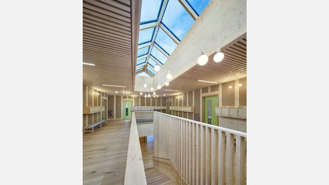 Lichthof: Galerie mit Holzbrüstung, großes Oberlicht und runde Pendelleuchten; grüne Türen zu Nebenräumen.
