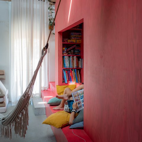 „Ein heller Innenraum mit Holzdecke und einer markanten, rosaroten Wand. Darin integriert ist eine gemütliche Leseecke mit Büchern und bunten Kissen, davor hängt eine helle Hängematte.“