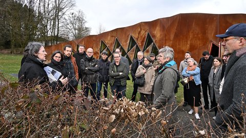 Gruppe von Menschen steht vor einer rostbraunen, wellenförmigen Wand mit dreieckigen Fenstern und hört einer Frau zu, die Papiere hält