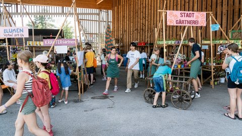 "Inneren einer großen Holzscheune mit hohem Dach und großem offenen Tor. Mehrere Kinder und Jugendliche versammeln sich in Gruppen. Schilder mit Aufschriften wie 'STADT GÄRTNEREI' und 'KRÄUTER Gärtner' hängen an Holzgestellen. Pflanzen in Blumentöpfen und alte Gegenstände sind ausgestellt. Ein Mädchen fährt Fahrrad."
