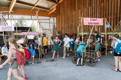 "Inneren einer großen Holzscheune mit hohem Dach und großem offenen Tor. Mehrere Kinder und Jugendliche versammeln sich in Gruppen. Schilder mit Aufschriften wie 'STADT GÄRTNEREI' und 'KRÄUTER Gärtner' hängen an Holzgestellen. Pflanzen in Blumentöpfen und alte Gegenstände sind ausgestellt. Ein Mädchen fährt Fahrrad."