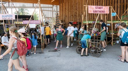 "Inneren einer großen Holzscheune mit hohem Dach und großem offenen Tor. Mehrere Kinder und Jugendliche versammeln sich in Gruppen. Schilder mit Aufschriften wie 'STADT GÄRTNEREI' und 'KRÄUTER Gärtner' hängen an Holzgestellen. Pflanzen in Blumentöpfen und alte Gegenstände sind ausgestellt. Ein Mädchen fährt Fahrrad."