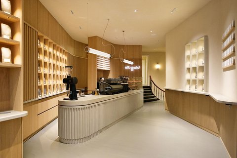 „Helles, modern eingerichtetes Café der Marke ‚elbgold‘. Im Zentrum steht ein langer, geriffelter heller Tresen mit einer schwarzen Espressomaschine. Die Wände sind mit hellem Holz verkleidet und in die raumhohen Regale sind Kaffeetüten einsortiert. Über dem Tresen hängen minimalistische, röhrenförmige Lampen.“