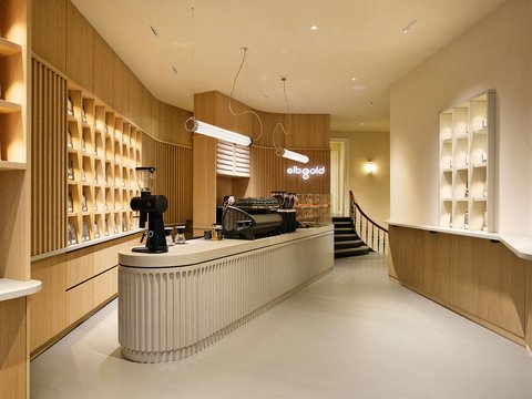 „Helles, modern eingerichtetes Café der Marke ‚elbgold‘. Im Zentrum steht ein langer, geriffelter heller Tresen mit einer schwarzen Espressomaschine. Die Wände sind mit hellem Holz verkleidet und in die raumhohen Regale sind Kaffeetüten einsortiert. Über dem Tresen hängen minimalistische, röhrenförmige Lampen.“