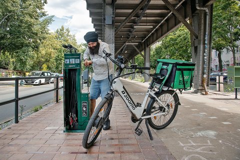 Mann mit Turban und Bart lädt E-Bike an Ladestation in urbaner Umgebung auf