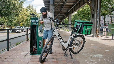 Mann mit Turban und Bart lädt E-Bike an Ladestation in urbaner Umgebung auf