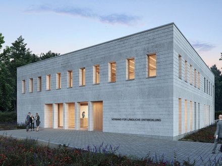 Außenansicht: Zweigeschossiger, heller Mauerwerksbau in Dämmerung; schmale Fenster mit warmem Licht, gepflasterter Vorplatz und Wildblumen; an der Fassade steht „Verband für ländliche Entwicklung“.