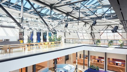 Großzügiges Atrium mit Glasdach, Stahlkonstruktion und Blick auf Sitz- und Arbeitsbereiche über mehrere Ebenen.