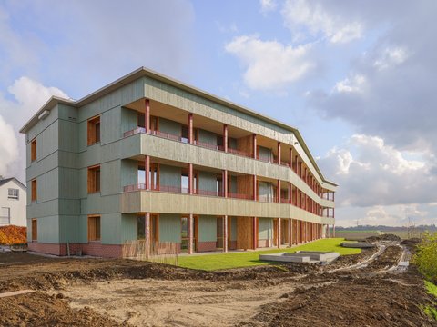 Dreigeschossiges Wohnhaus mit durchgehenden Balkonen, grüner Holzfassade und Klinkersockel; Außenanlagen noch im Bau mit Erdflächen und frischem Rasen.