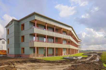 Dreigeschossiges Wohnhaus mit durchgehenden Balkonen, grüner Holzfassade und Klinkersockel; Außenanlagen noch im Bau mit Erdflächen und frischem Rasen.