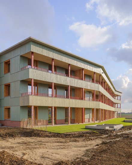 Dreigeschossiges Wohnhaus mit durchgehenden Balkonen, grüner Holzfassade und Klinkersockel; Außenanlagen noch im Bau mit Erdflächen und frischem Rasen.