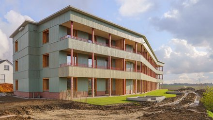 Dreigeschossiges Wohnhaus mit durchgehenden Balkonen, grüner Holzfassade und Klinkersockel; Außenanlagen noch im Bau mit Erdflächen und frischem Rasen.