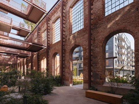 "Innenhof einer ehemaligen Fabrik mit massiven roten Backsteinmauern und großen Spitzbogenöffnungen. Durch die Bögen sind moderne Wohngebäude im Hintergrund sichtbar. Der Hof ist mit Grün bepflanzt, mehrere Holzbänke laden zum Verweilen ein. Das Dach des Innenhofs ist teilweise verglast. Eine nachhaltige Umnutzung einer historischen Industrieanlage."