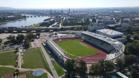 Luftaufnahme eines modernen Leichtathletikstadions mit markantem weißen Dach am Flussufer, im Hintergrund eine historische Stadtsilhouette.