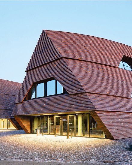 Moderne Schulgebäude mit geneigten Fassaden aus dunklen Holzschindeln auf offenem Platz.