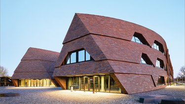 Moderne Schulgebäude mit geneigten Fassaden aus dunklen Holzschindeln auf offenem Platz.