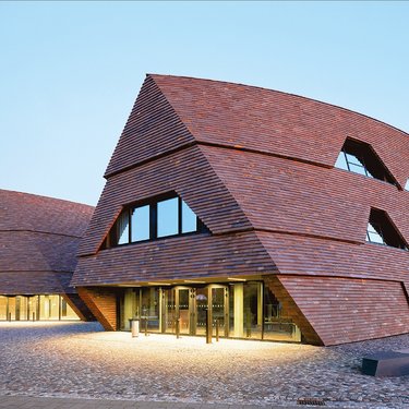 Moderne Schulgebäude mit geneigten Fassaden aus dunklen Holzschindeln auf offenem Platz.