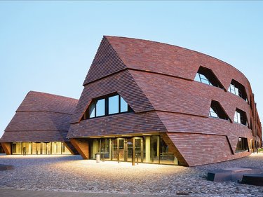 Moderne Schulgebäude mit geneigten Fassaden aus dunklen Holzschindeln auf offenem Platz.