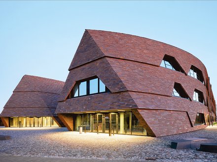 Moderne Schulgebäude mit geneigten Fassaden aus dunklen Holzschindeln auf offenem Platz.