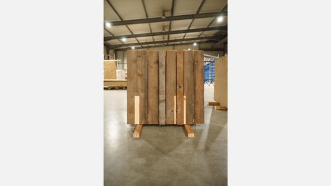 Freistehendes Musterstück: Wand aus verwitterten Holzbohlen mit hellen Einlagen in einer Werkhalle.