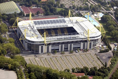 Schrägaufnahme aus der Luft auf den Signal Iduna Park in Dortmund. Das Stadion besticht durch seine markanten, leuchtend gelben Pylone, die das Dach tragen. Die Dachflächen sind teilweise mit Solarmodulen bedeckt. Der Schriftzug "SIGNAL IDUNA PARK" ist gut sichtbar auf der Haupttribüne angebracht. Das Stadion ist umgeben von Parkplätzen, Bäumen, einem Freibad im Hintergrund und einer Laufbahn.