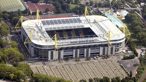 Schrägaufnahme aus der Luft auf den Signal Iduna Park in Dortmund. Das Stadion besticht durch seine markanten, leuchtend gelben Pylone, die das Dach tragen. Die Dachflächen sind teilweise mit Solarmodulen bedeckt. Der Schriftzug "SIGNAL IDUNA PARK" ist gut sichtbar auf der Haupttribüne angebracht. Das Stadion ist umgeben von Parkplätzen, Bäumen, einem Freibad im Hintergrund und einer Laufbahn.