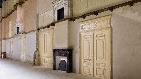 "Großflächige Architektur-Installation mit Rekonstruktionen historischer Wohnraumelemente. Links steht eine rosa getönte Wand mit weißen Türen und Treppenlauf aus Marmor. Rechts eine olivgelbe Wand mit weißen Türelementen und Details. Zentral befindet sich eine schwarze Vintage-Kaminumrandung aus Marmor. Die Installation zeigt die Kontraste verschiedener klassischer Stilelemente. Helle, fokussierte Beleuchtung hebt die Architektur-Details hervor. Der weiße Betonboden bildet die Grundfläche."