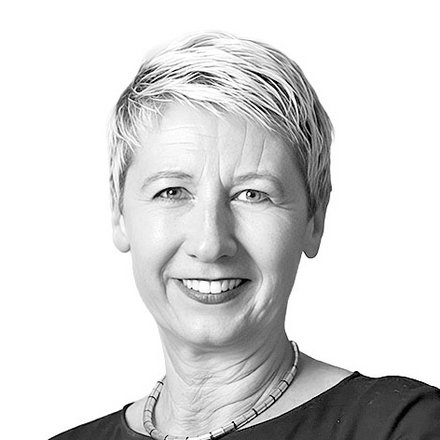 Schwarz-Weiß-Porträt einer Frau mit kurzen, hellgrauen Haaren, die direkt in die Kamera lächelt. Sie trägt ein schwarzes Shirt und eine silberfarbene Halskette. Der Hintergrund ist weiß.