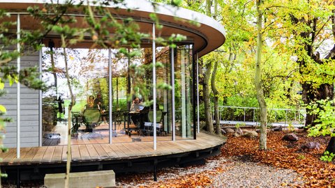 Runder Glaspavillon im herbstlichen Baumbestand; aufgeständert über Holzsteg und Kies, Einblicke ins Innere.