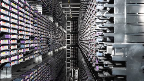 Man blickt in einen langen, engen Gang zwischen zwei hohen Wänden aus dicht gestapelten Servermodulen. Tausende identische Elemente erzeugen ein starkes Gefühl von Tiefe und Wiederholung. Das Bild macht die enorme Dichte und Materialität digitaler Infrastruktur deutlich. Es vermittelt Größe, Komplexität und Energieaufwand moderner Datenverarbeitung.