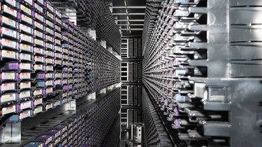 Man blickt in einen langen, engen Gang zwischen zwei hohen Wänden aus dicht gestapelten Servermodulen. Tausende identische Elemente erzeugen ein starkes Gefühl von Tiefe und Wiederholung. Das Bild macht die enorme Dichte und Materialität digitaler Infrastruktur deutlich. Es vermittelt Größe, Komplexität und Energieaufwand moderner Datenverarbeitung.