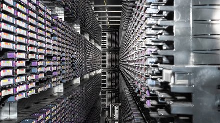 Man blickt in einen langen, engen Gang zwischen zwei hohen Wänden aus dicht gestapelten Servermodulen. Tausende identische Elemente erzeugen ein starkes Gefühl von Tiefe und Wiederholung. Das Bild macht die enorme Dichte und Materialität digitaler Infrastruktur deutlich. Es vermittelt Größe, Komplexität und Energieaufwand moderner Datenverarbeitung.