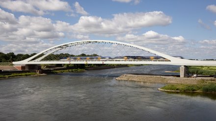 Seitenansicht einer weißen Eisenbahnbrücke mit Stahlbogen, ein Zug überquert den Fluss darunter.