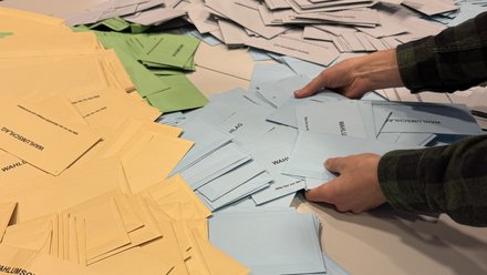 Zwei Hände die nach blauen, grünen und gelben Wahlscheinen greifen