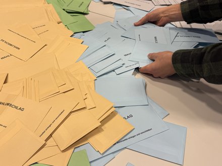 Zwei Hände die nach blauen, grünen und gelben Wahlscheinen greifen