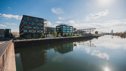 Uferpromenade mit modernen Bürogebäuden am Flussbecken; Glasfassaden spiegeln sich im Wasser, sonniger Himmel.