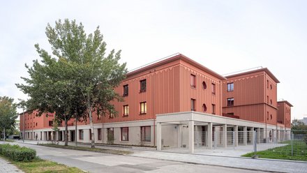 Langes, terrakottafarbenes Gebäude mit profilierter Fassade; Straßenbäume und überdachter Eingang an der Ecke.